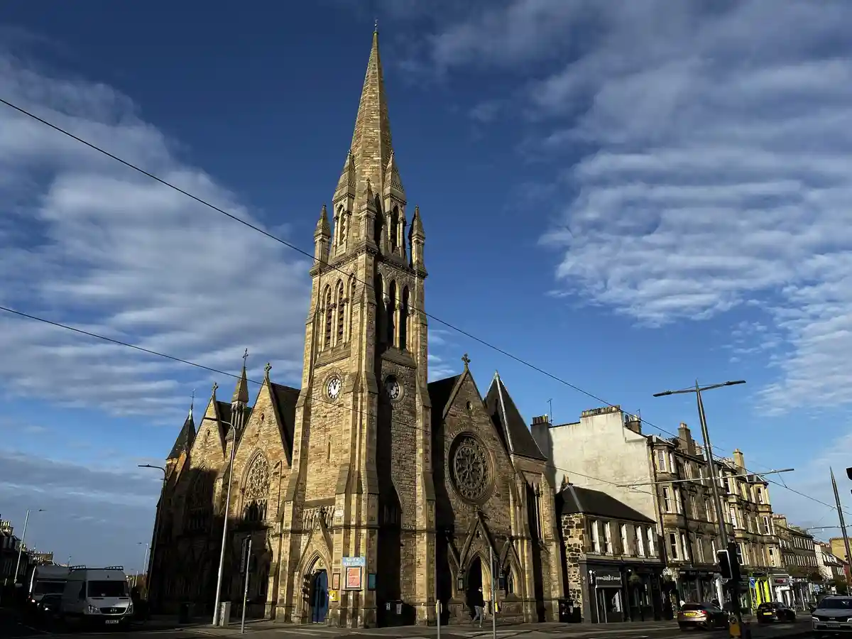 A grand, Gothic-style stone church with a tall spire and clock faces stands prominently against a blue sky with scattered…