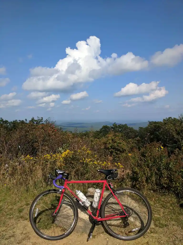 A red road bicycle with purple handlebars and two water bottles is parked on a dirt path atop a hill, overlooking a scenic…