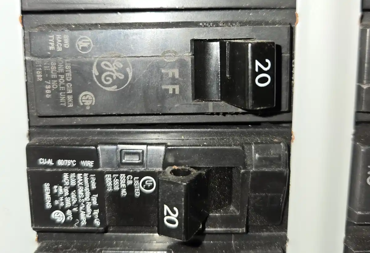 A close-up of two 20-amp circuit breakers in an electrical panel, one by GE and one by Siemens, both labeled '20' and both…