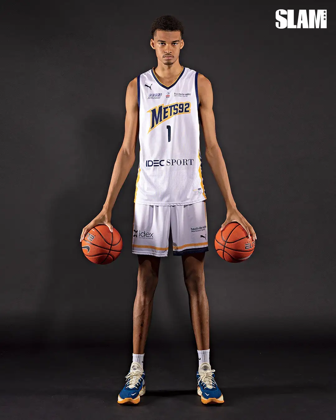 A tall young man in a white Mets92 basketball uniform with the number 1 stands against a dark background, holding one…