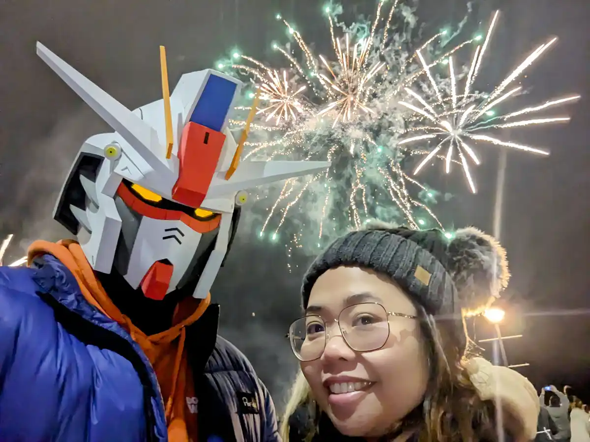 A selfie shows a person in a large white, red, and blue Gundam robot head costume standing next to a smiling woman wearing…