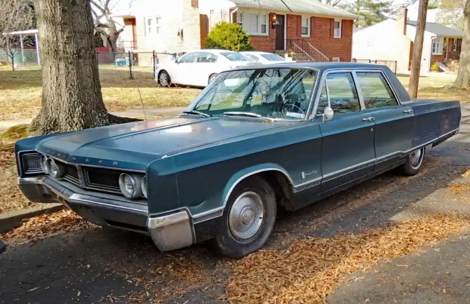 A teal 1960s Chrysler Imperial sedan is parked on a residential street covered in fallen autumn leaves. The car appears aged…