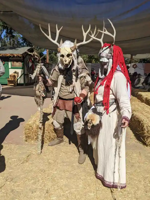 Two people in elaborate fantasy costumes stand side by side under a tent. The person on the left wears a deer skull mask…