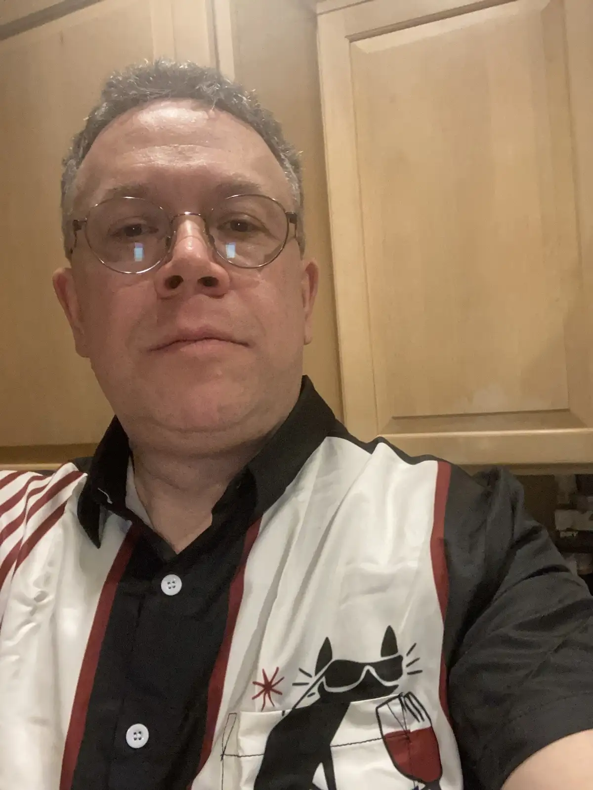 A middle-aged man with glasses and short graying hair takes a selfie in a kitchen, wearing a black shirt under a white and…