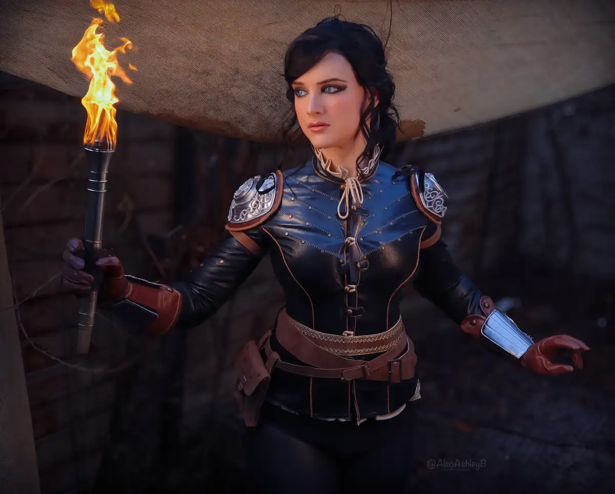 A person in a fantasy-themed costume, holding a flaming torch, looks off to the side with a neutral expression. The costume…