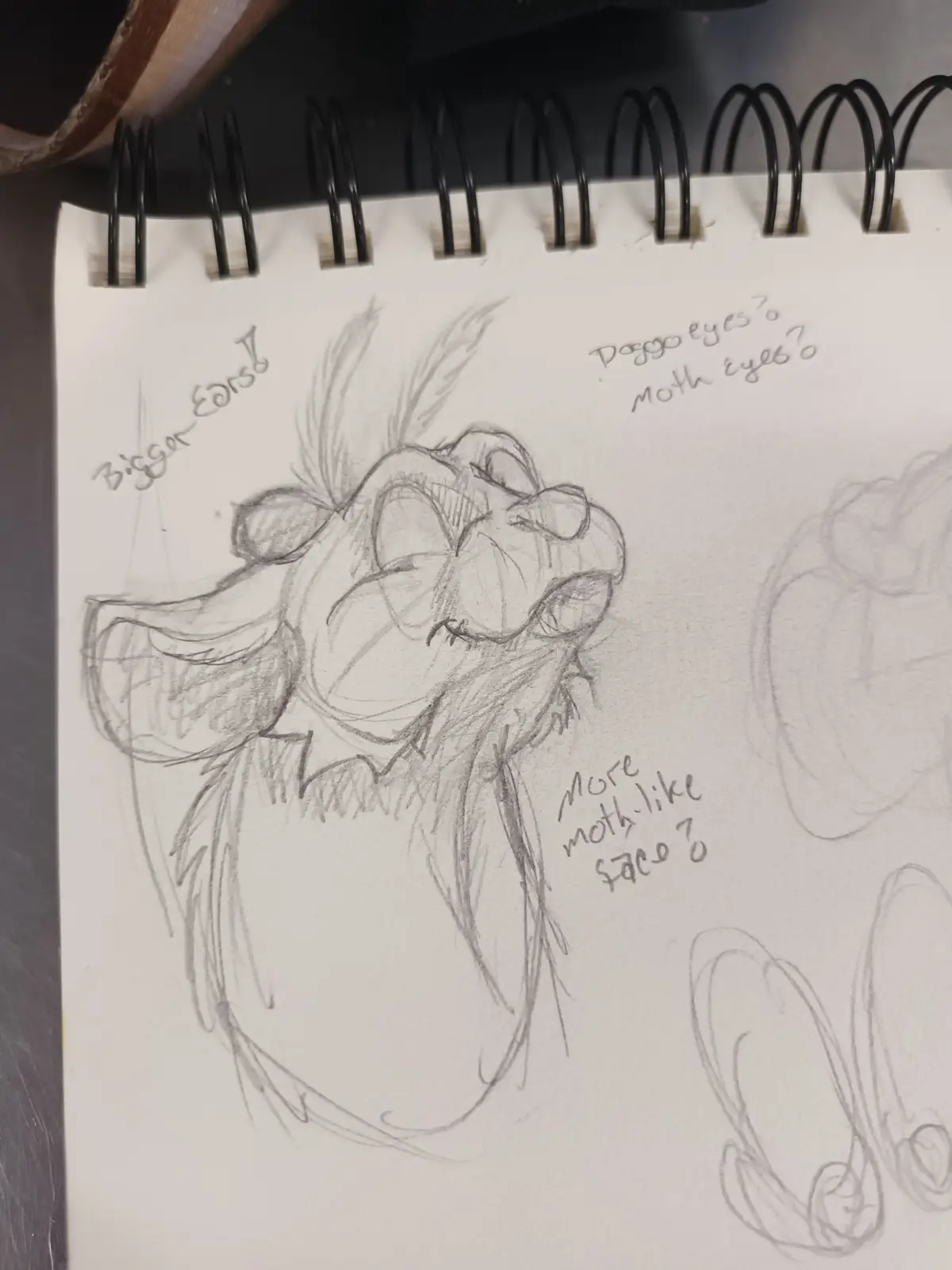 A pencil sketch in a spiral-bound notebook shows a cartoonish creature with large ears and expressive eyes, surrounded by…