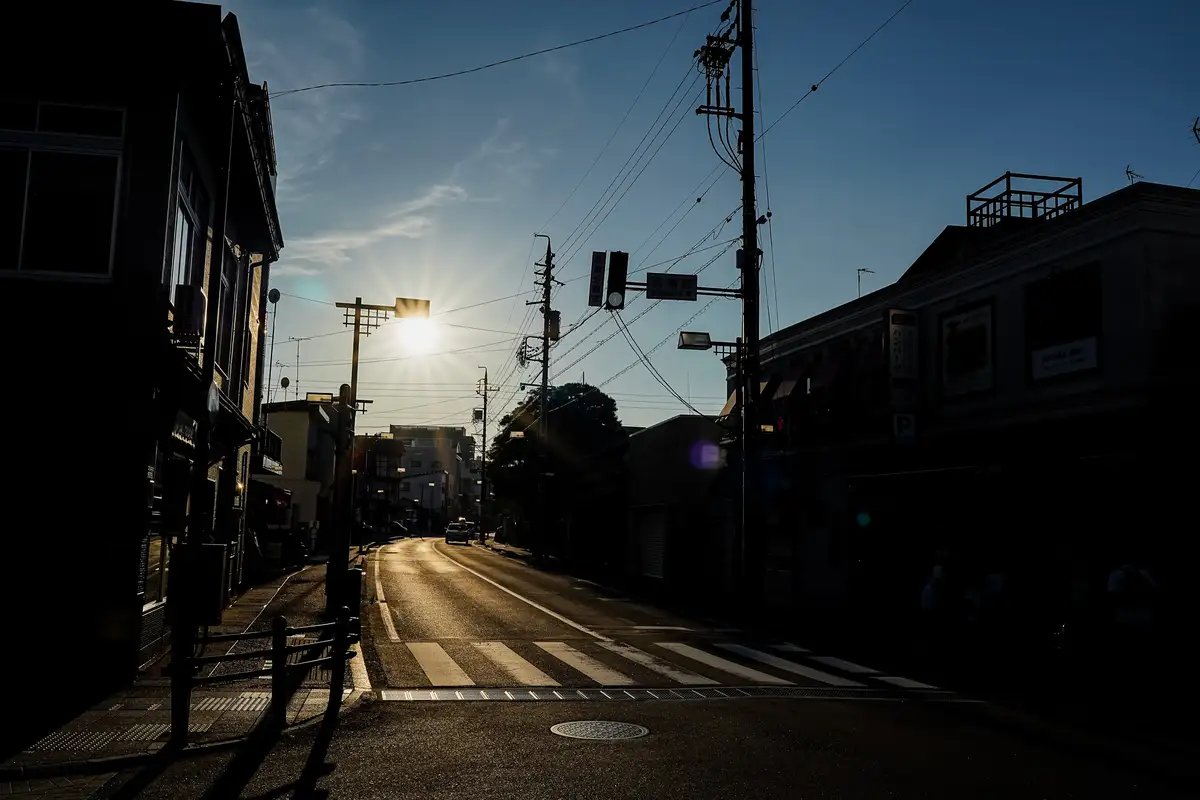 A low-angle shot of a quiet Japanese street at sunrise or sunset, with the sun creating a bright starburst effect in the…