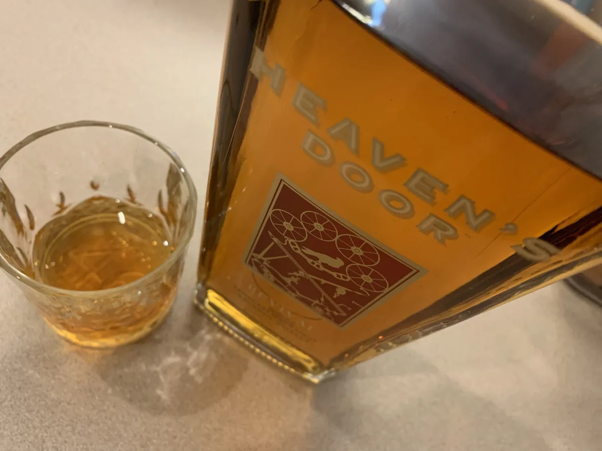 A close-up photo shows a bottle of Heaven's Door whiskey next to a glass containing amber liquid, both resting on a…