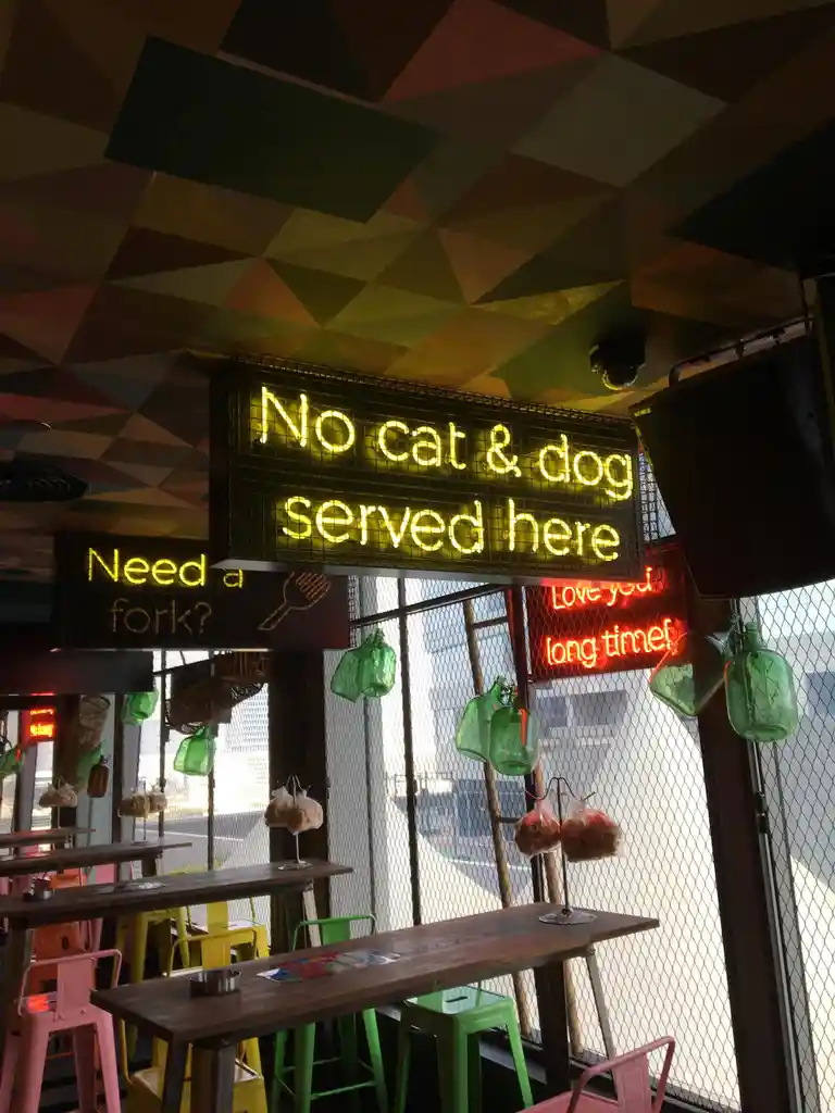 A photo of a restaurant interior with colorful geometric ceiling tiles, wooden tables, and bright green, pink, and yellow…