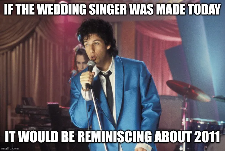 A man in a blue suit and black tie holds a microphone, singing with an intense expression. Behind him, a woman is partially…