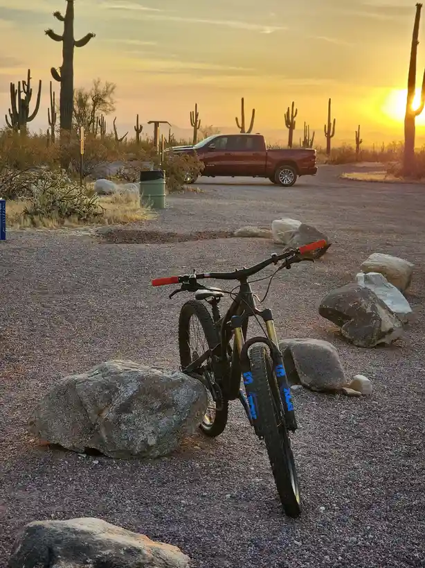 A mountain bike with red handlebars and blue Fox suspension forks is parked on gravel next to a large rock in a desert…