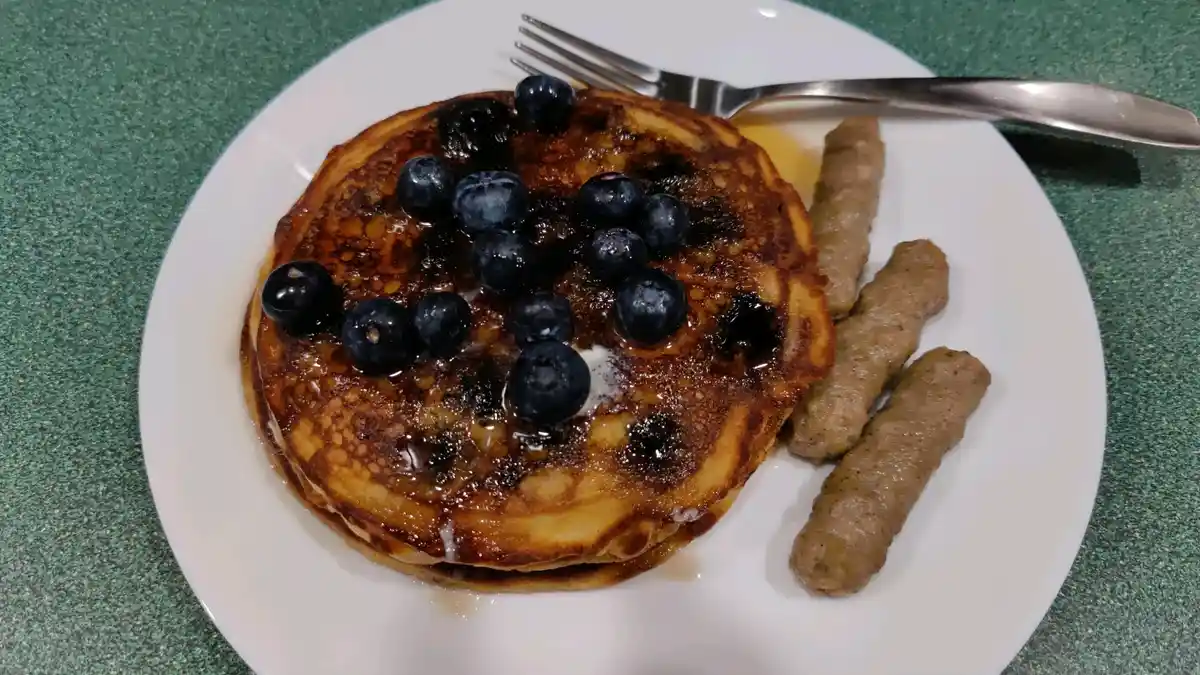 A close-up photo shows a white plate with a stack of golden-brown pancakes topped with syrup and fresh blueberries…
