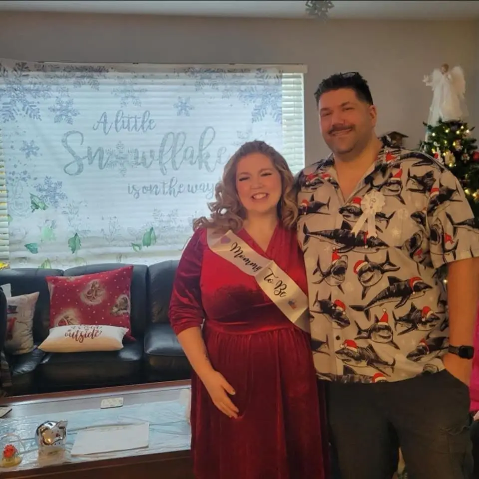 A pregnant woman in a red dress wearing a 'Mommy To Be' sash stands beside a man in a shark-print shirt with Santa hats…