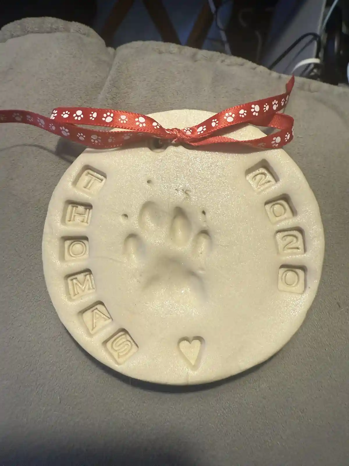 A circular, off-white ceramic keepsake with a raised paw print in the center, surrounded by the name 'THOMAS' on the left…