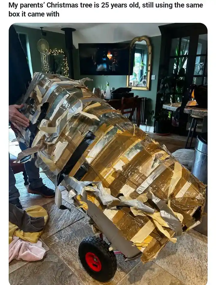 A person is holding a large, heavily taped cardboard box that appears to be a Christmas tree stand or base, covered in…