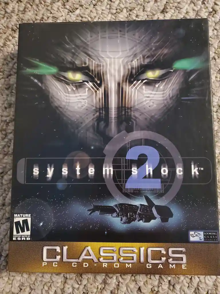The image shows the cover art for the PC game 'System Shock 2', featuring a menacing, circuit-board-like face with glowing…