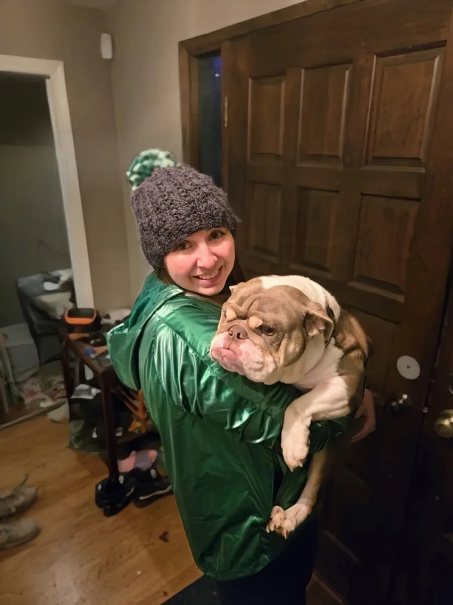 A person wearing a shiny green jacket and a grey knit hat with a green pom-pom is smiling while holding a bulldog. The dog…