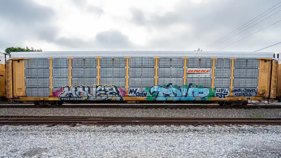 A side view of a BNSF freight train car covered in colorful graffiti tags, sitting on railway tracks under a cloudy sky. The…