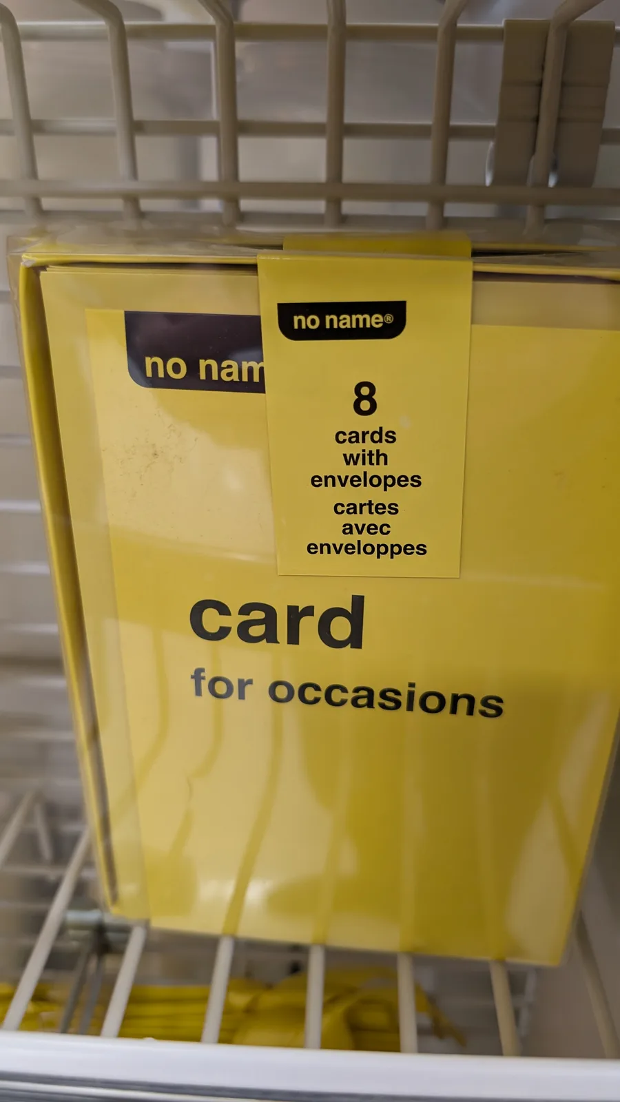 A yellow box of 'no name' brand greeting cards sits on a store shelf, labeled '8 cards with envelopes' and 'card for…