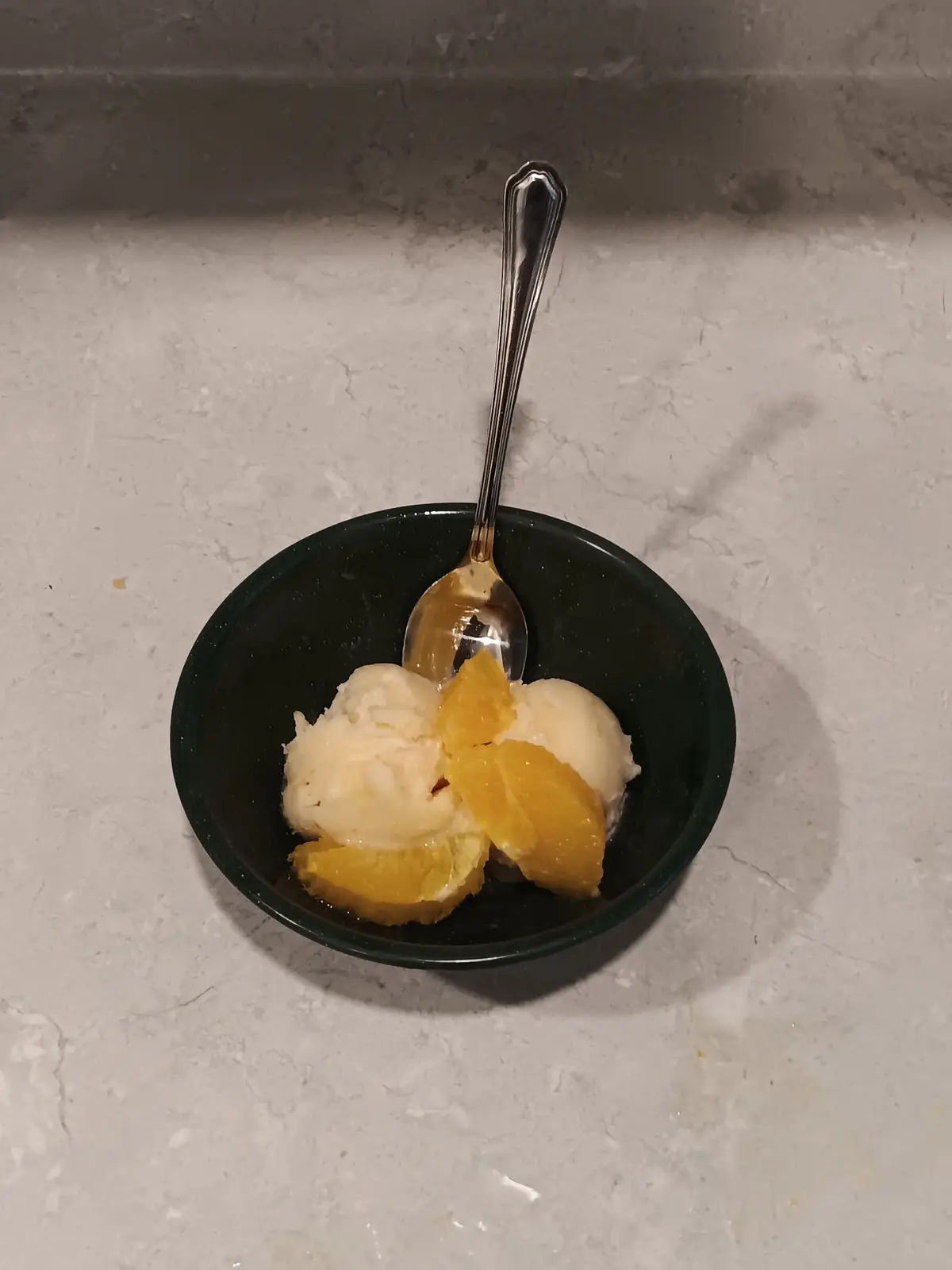 A dark green bowl sits on a light gray marble countertop, containing two scoops of vanilla ice cream and several orange…