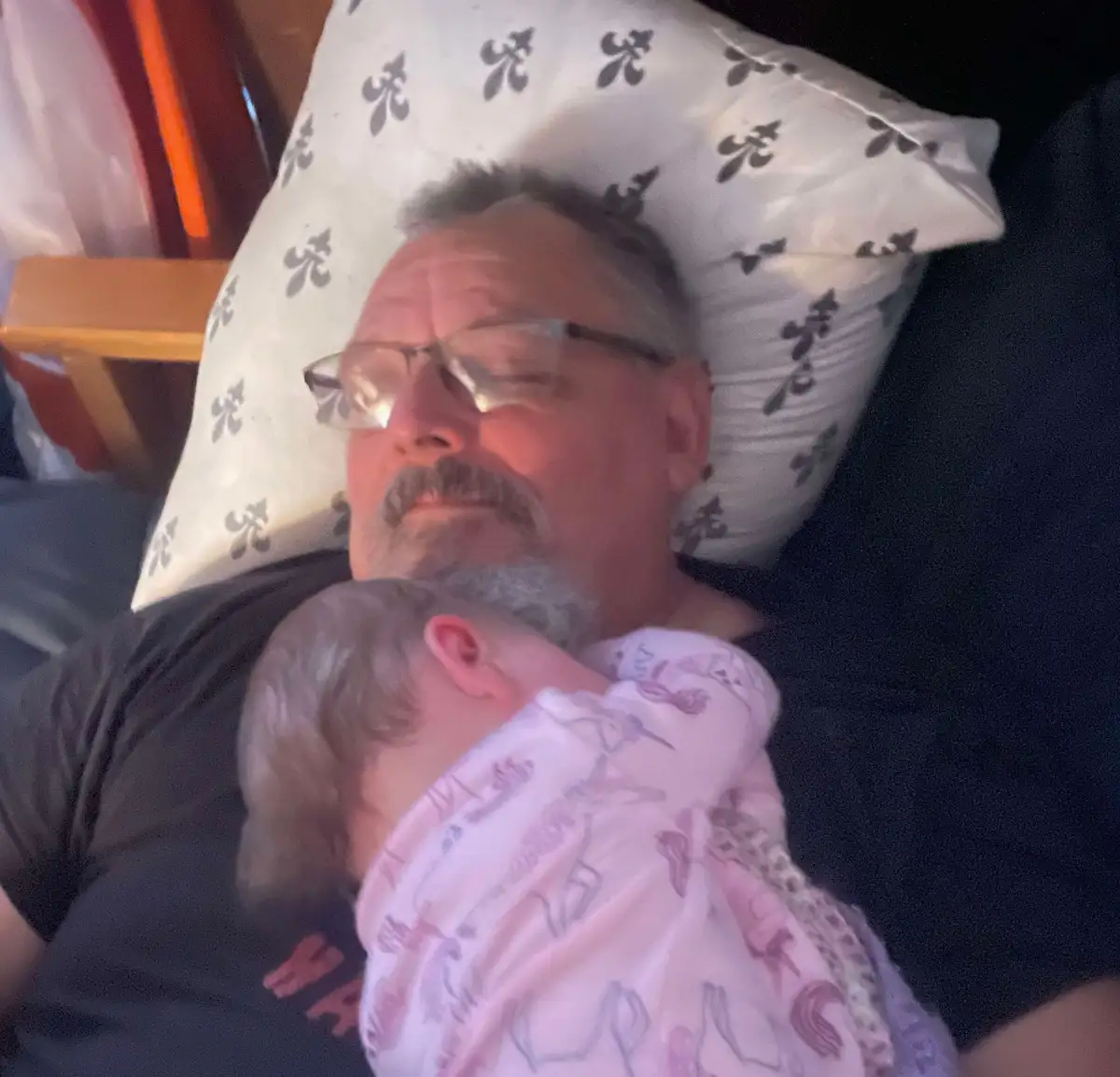 An older man with glasses and a mustache is sleeping on a patterned pillow while holding a baby wrapped in a pink onesie…