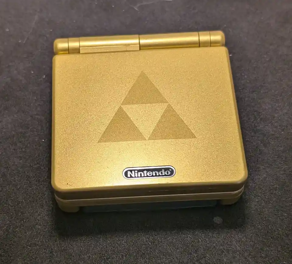 A gold-colored Nintendo Game Boy Advance is shown closed, resting on a dark gray surface. The device features the iconic…