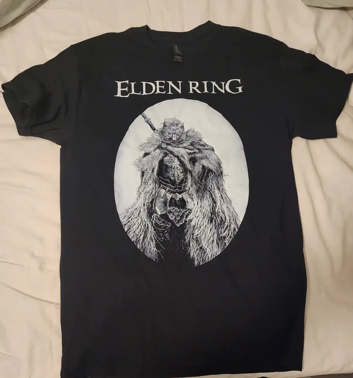 A black t-shirt laid flat on a bed, featuring the text 'ELDEN RING' in a stylized font above a circular illustration of a…