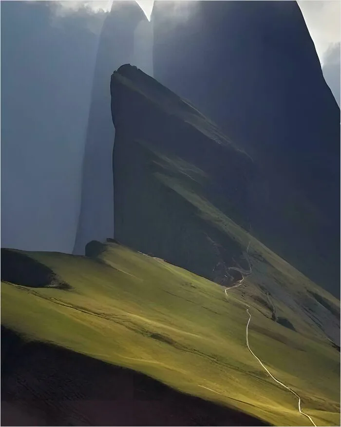 A dramatic landscape photo shows a winding dirt path ascending a steep, grassy hill toward towering, shadowed rock…
