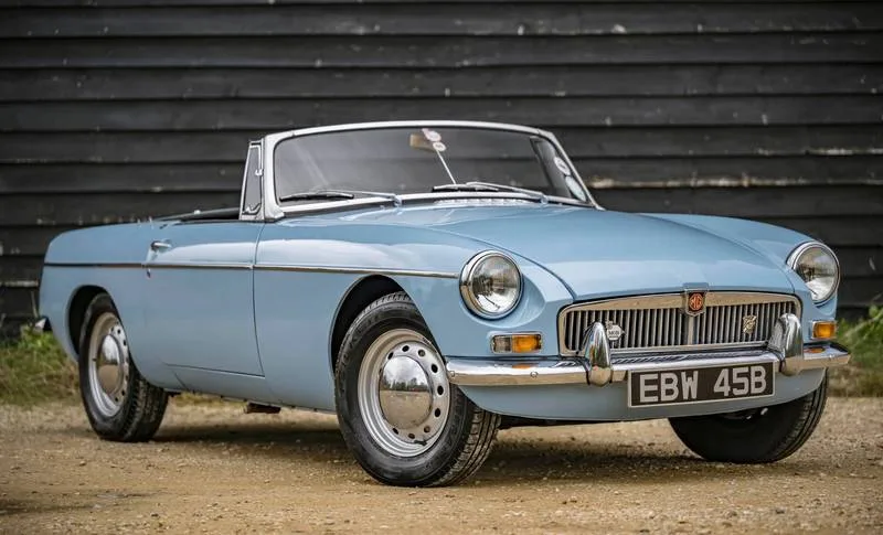 A light blue classic convertible car, likely an MG MGB, is parked on dirt in front of a dark wooden wall. The car has chrome…