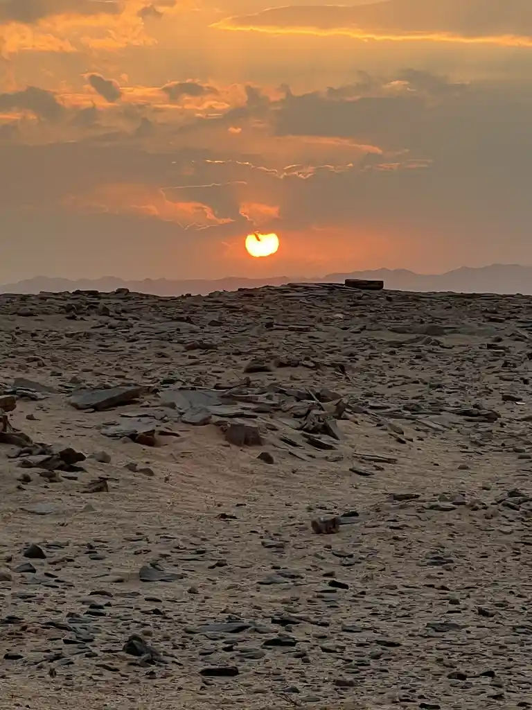 A scenic desert sunset with a bright orange sun partially obscured by clouds, casting warm light over a rocky, sandy terrain…