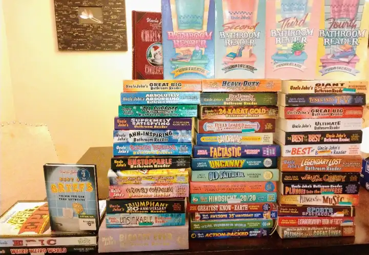 A large collection of Uncle John's Bathroom Reader books stacked in multiple piles on a shelf, with titles like 'Giant…