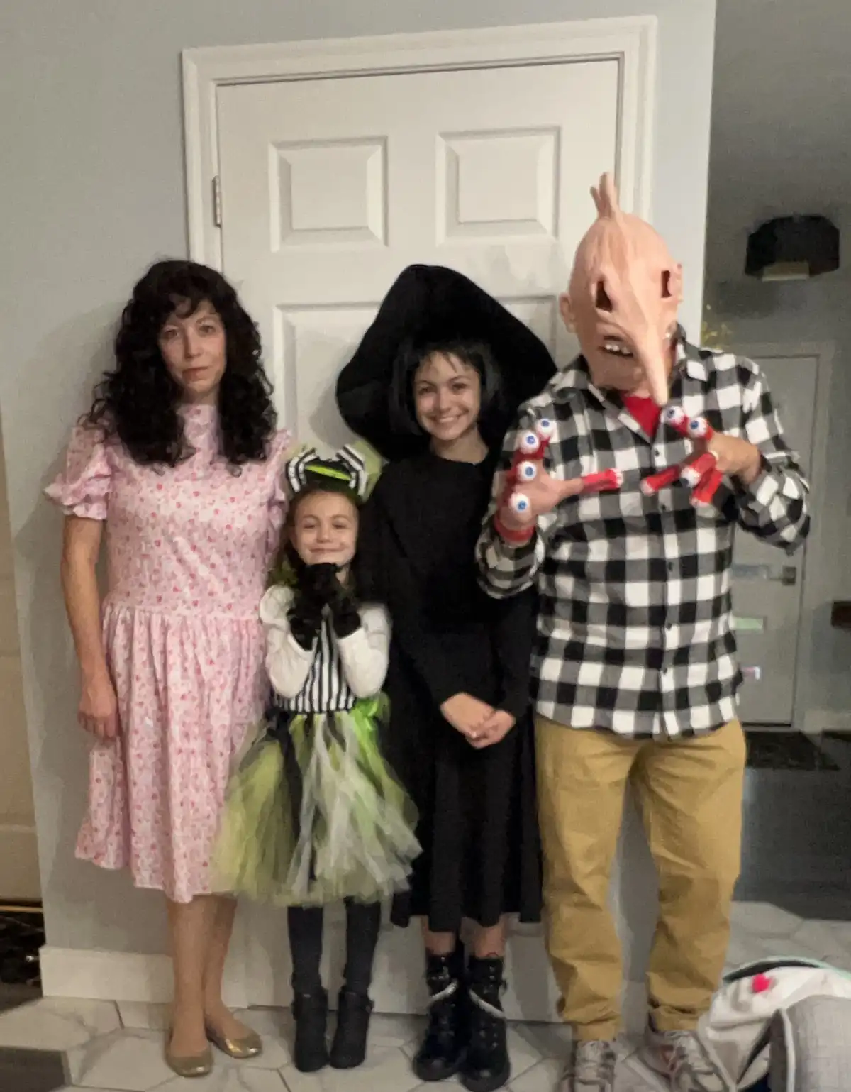 Four people pose indoors for a Halloween photo. From left to right: a woman in a pink floral dress, a young girl in a green…