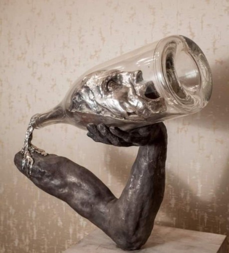 A dark-toned sculpture depicts a human arm and hand holding an oversized, tilted glass bottle. Inside the bottle, a…