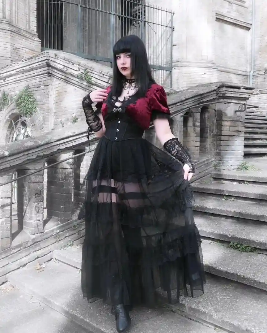 A person with black hair and bangs, wearing a red velvet top, black corset, and a layered black lace and tulle skirt, stands…