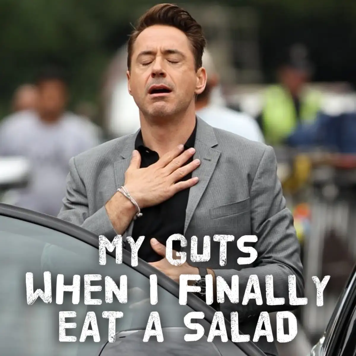 A man in a gray suit with his eyes closed and hand on his chest, standing next to a car, with the text 'MY GUTS WHEN I…