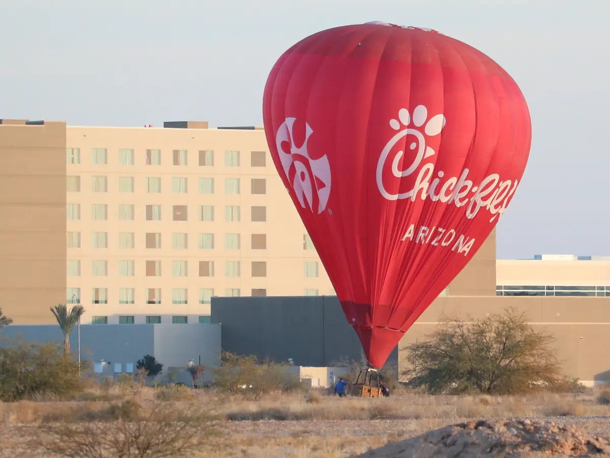 A large red hot air balloon with the Chick-fil-A logo and text 'Chick-fil-A ARIZONA' is shown on the ground in a desert-like…