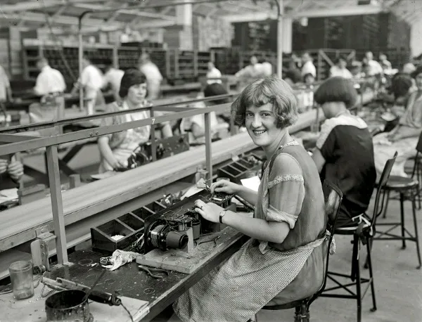 A vintage black-and-white photo shows a smiling woman in a factory setting, seated at a workbench with tools and machinery…