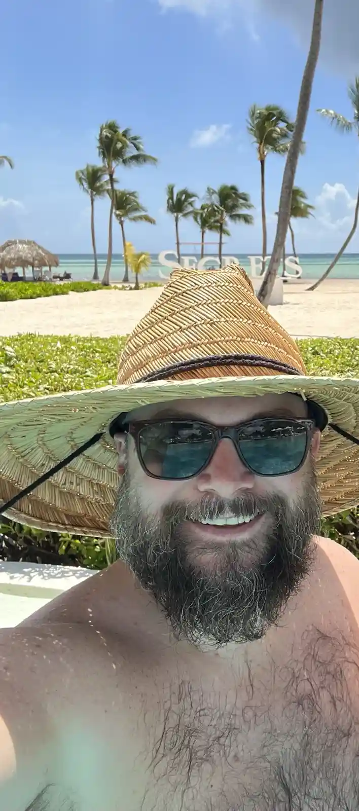 A man with a beard and sunglasses wearing a large straw hat smiles at the camera while shirtless on a beach. Behind him…
