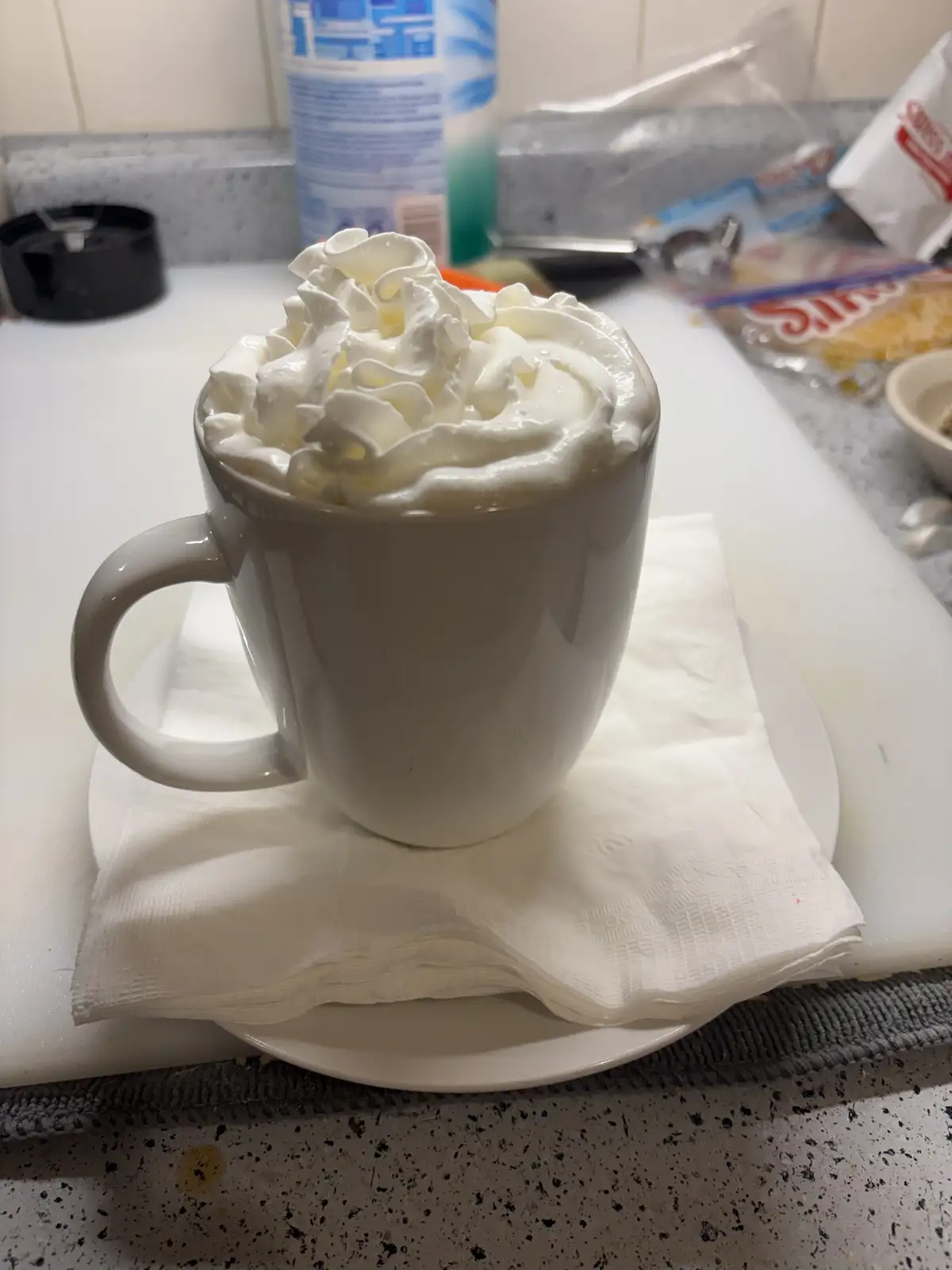 A white ceramic mug overflowing with whipped cream sits on a white napkin atop a saucer, placed on a kitchen counter. The…