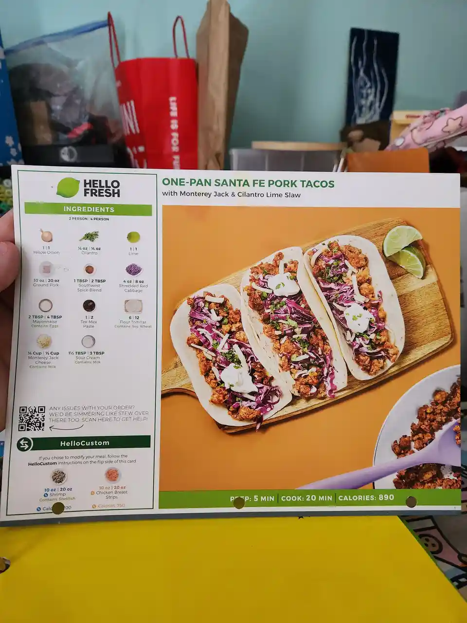 A person holds a HelloFresh recipe card for 'One-Pan Santa Fe Pork Tacos' showing ingredients and a photo of three tacos…