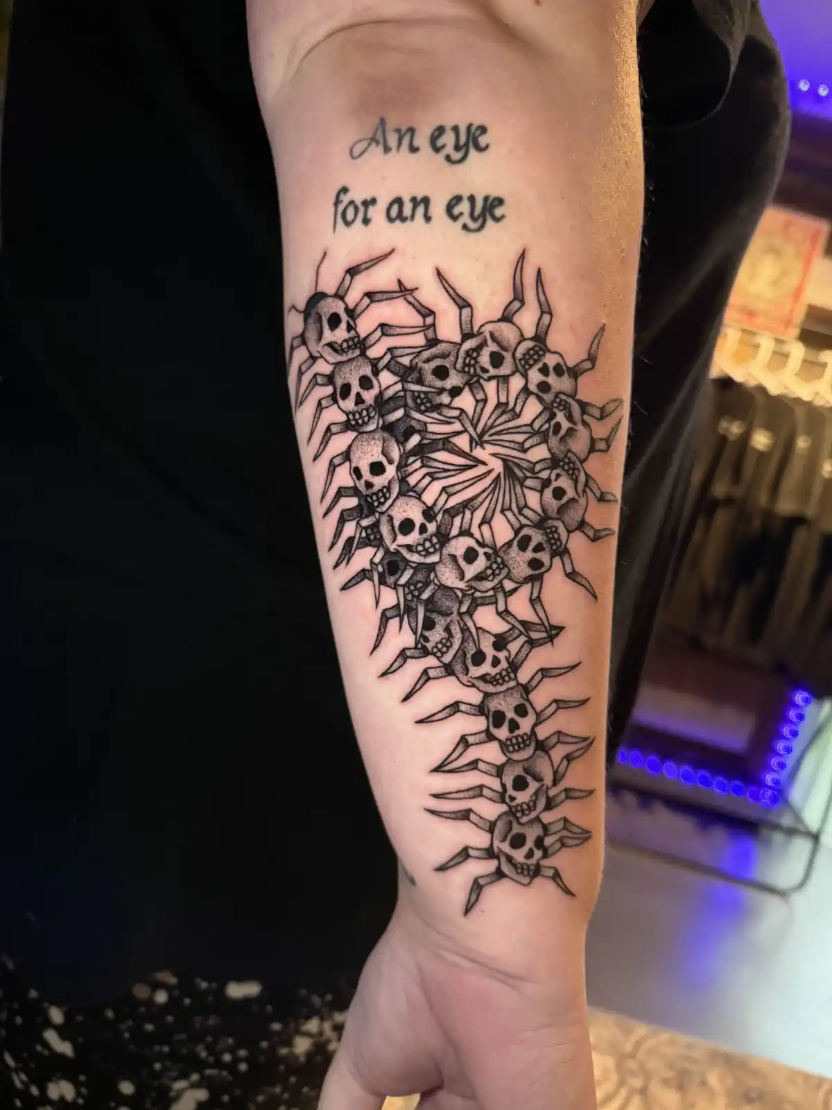 A person's forearm displays a black and grey tattoo featuring multiple skulls with spider-like legs arranged in a circular…