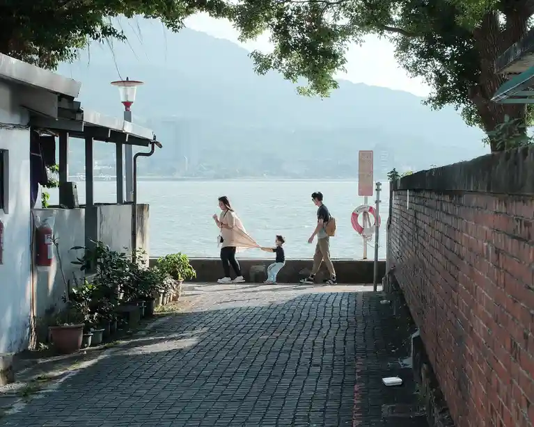 A family walks along a cobblestone path beside a body of water, with a child pulling on a woman's flowing dress. A red and…