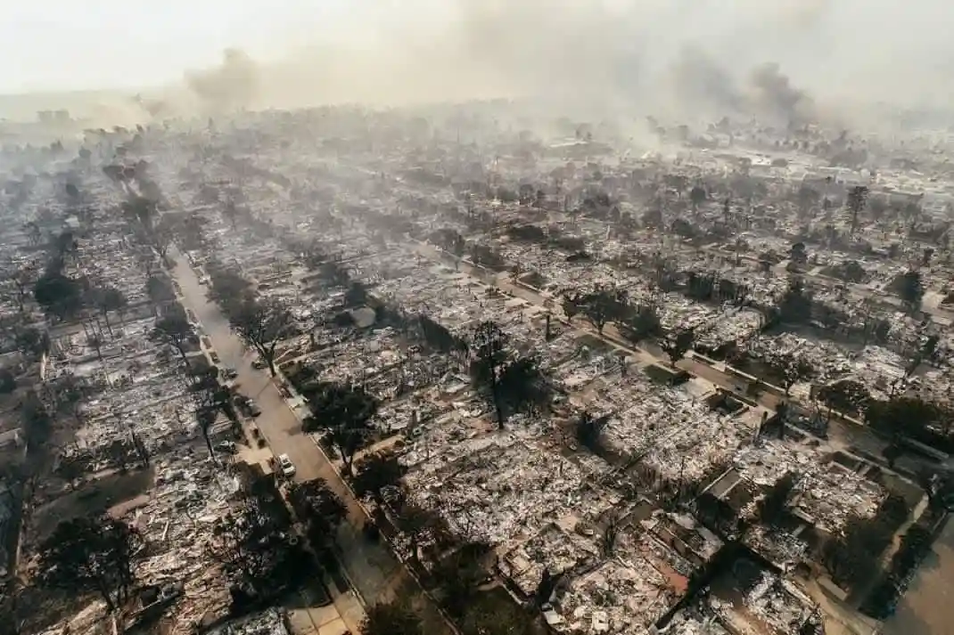 An aerial photograph shows a residential neighborhood completely destroyed by fire, with charred remains of houses…