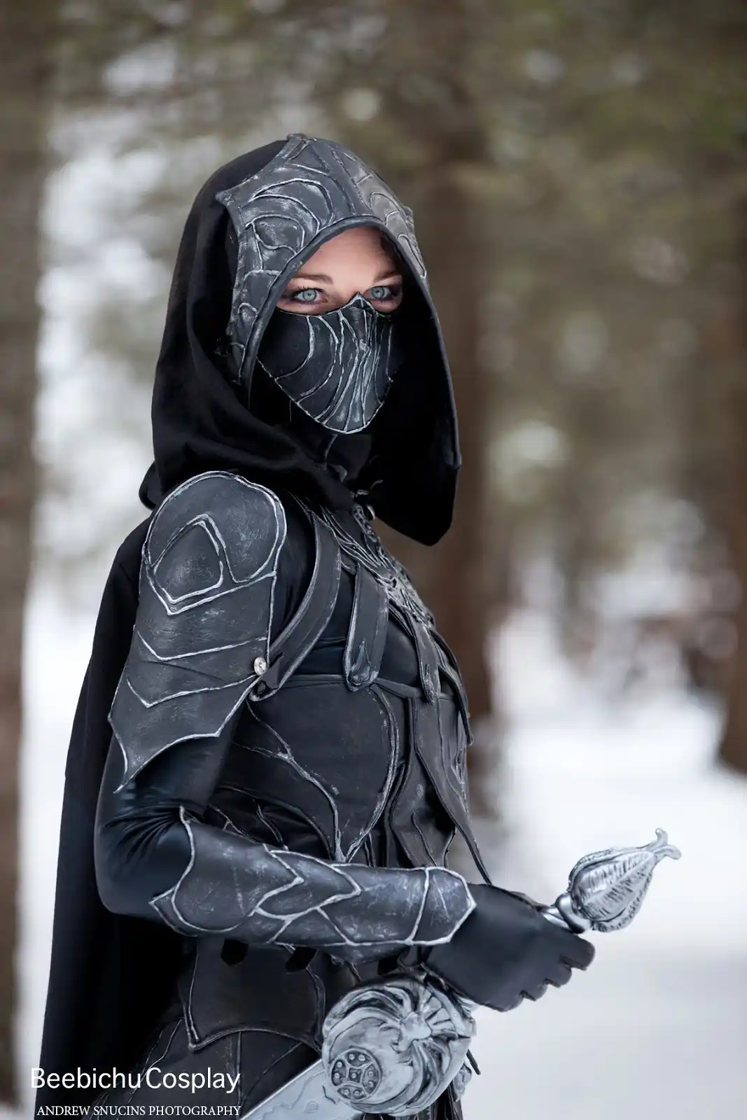 A person in a detailed black and silver armored cosplay costume with a hood and face mask, standing in a snowy forest while…