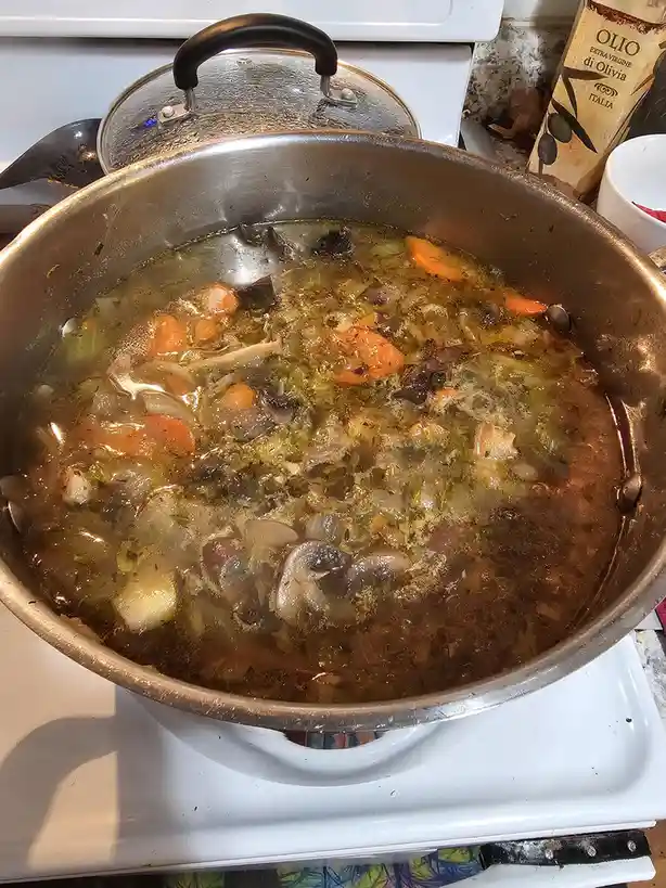 A large stainless steel pot sits on a white stovetop, filled with a murky, bubbling broth containing visible chunks of…