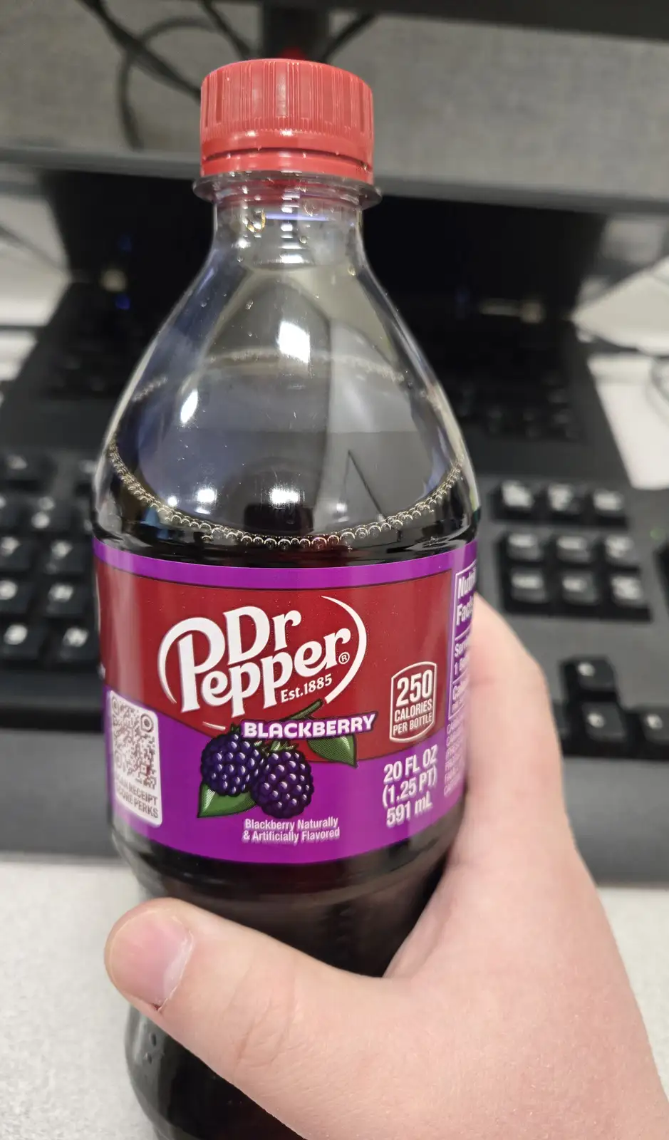 A hand holds a nearly empty Dr Pepper Blackberry soda bottle with a red cap and purple label, showing the brand name…