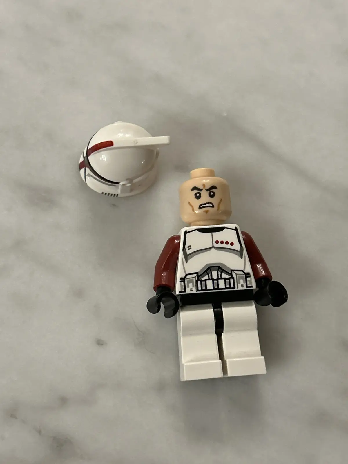 A LEGO Star Wars clone trooper minifigure stands on a marble surface with its white helmet lying nearby. The figure has an…