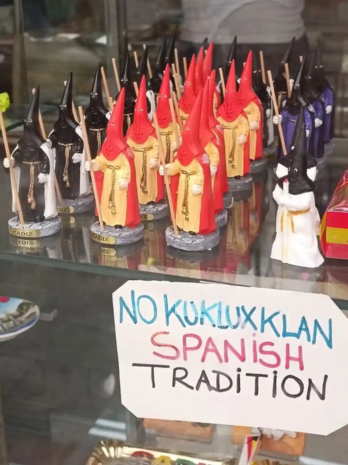 A shop window displays rows of small figurines dressed as Spanish 'Nazarenos' from Holy Week processions, wearing red or…