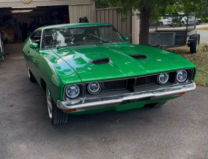 A shiny green classic muscle car, likely a Holden Monaro, is parked in front of a garage. The car has a distinctive hood…