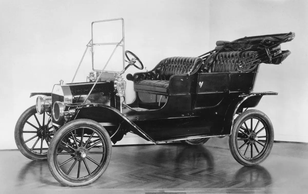 a black-and-white photograph shows a vintage 1909 Ford Model T automobile, featuring a tufted leather couch installed in the…